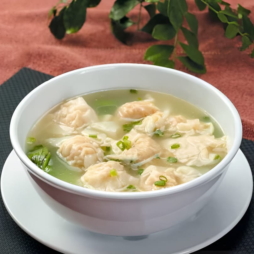 A white bowl of Bo Loong wonton soup with green onions and bok choy on a dark placemat.
