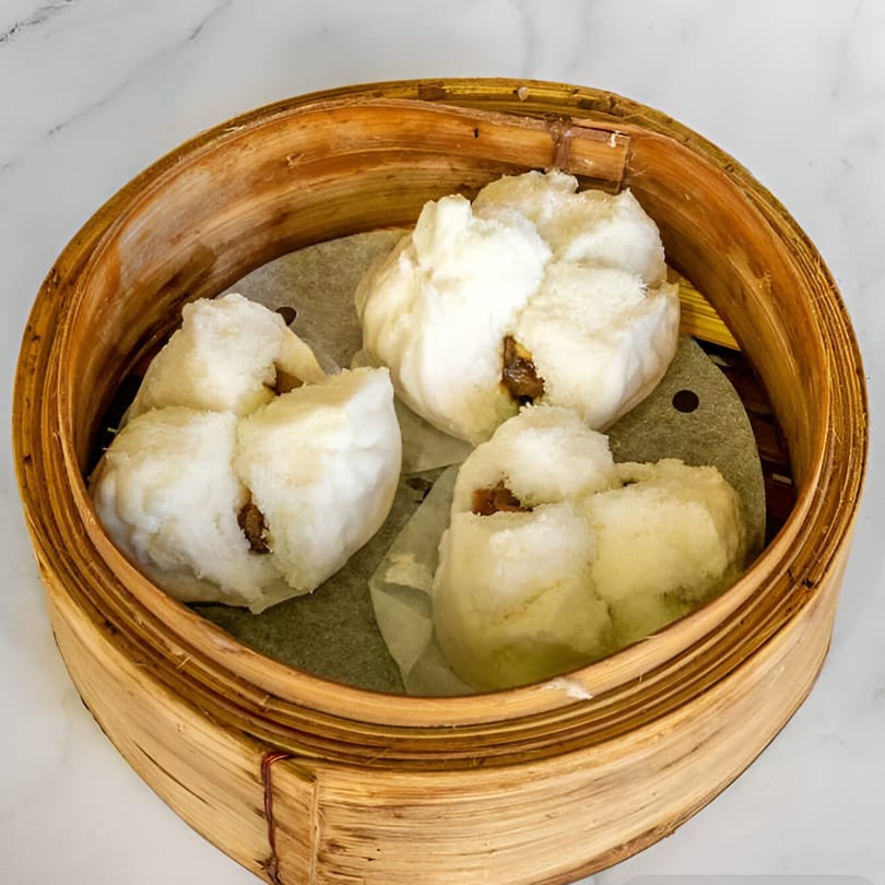 Three fluffy white char siu bao, or Chinese BBQ pork buns, in a bamboo steamer.